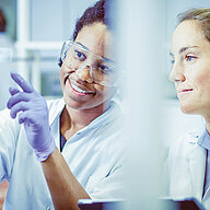 Two people in laboratory coats reviewing information together, with one person pointing off-screen while wearing protective safety glasses and gloves.