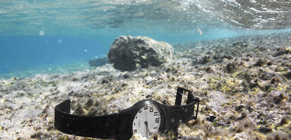 A watch with an analog dial and black plastic strap lies underwater on a stony surface