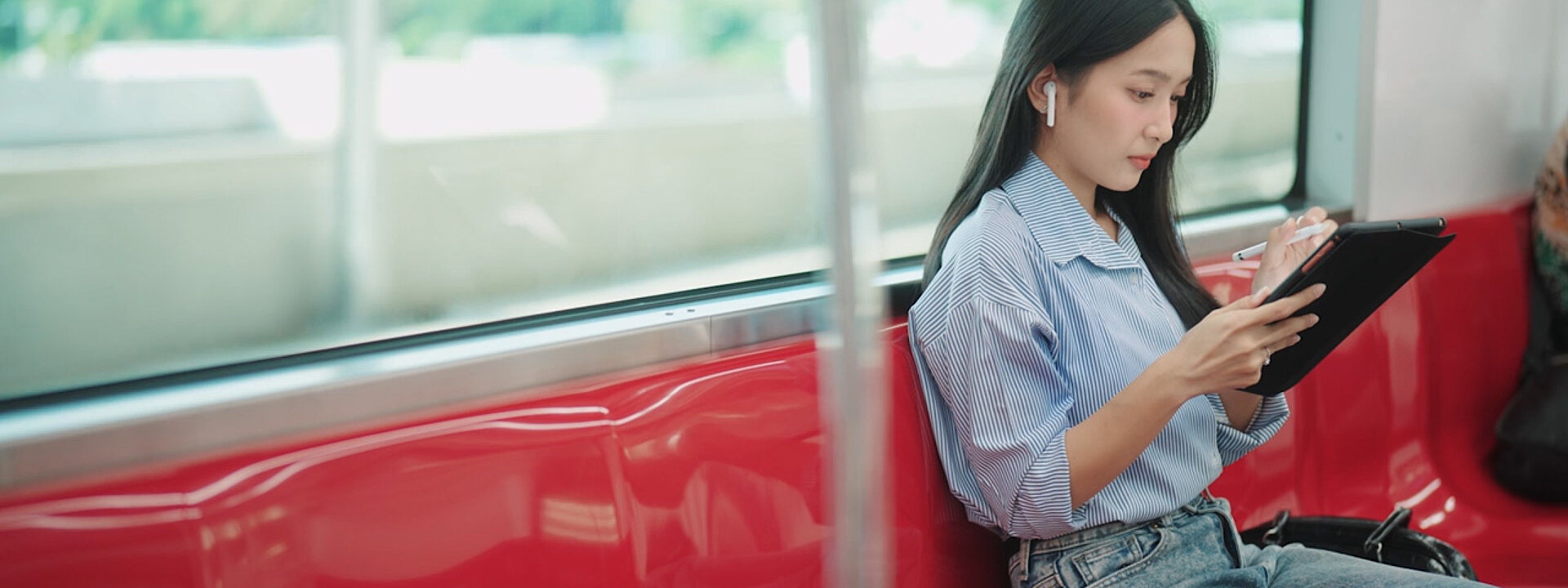 A woman sitting on a train with her back to the window. She is wearing earphones while working on her wireless device