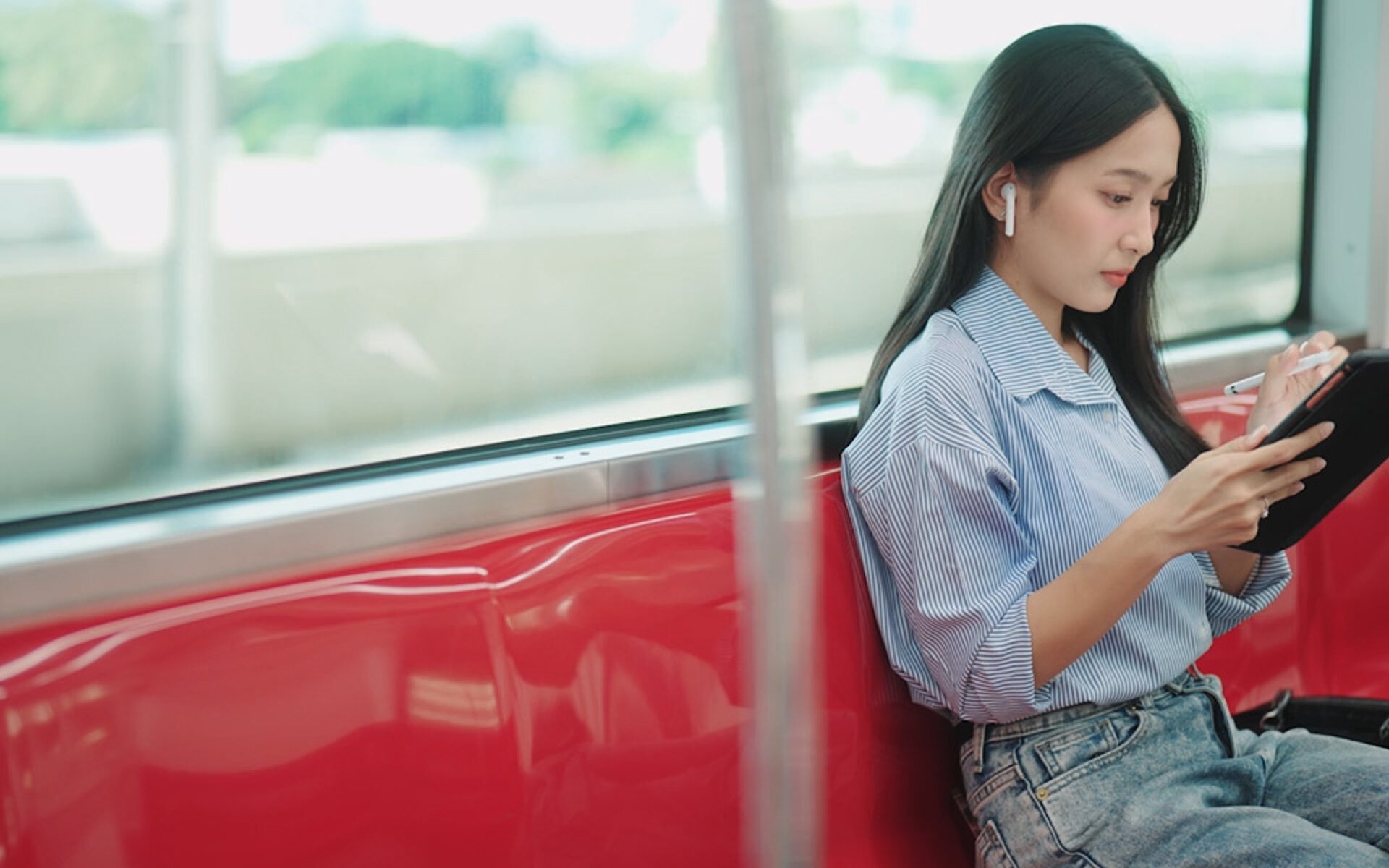 A woman sitting on a train with her back to the window. She is wearing earphones while working on her wireless device